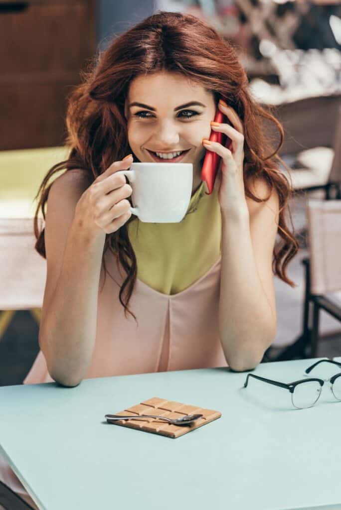 A smiling redhead woman with cup of coffee talking on smartphone